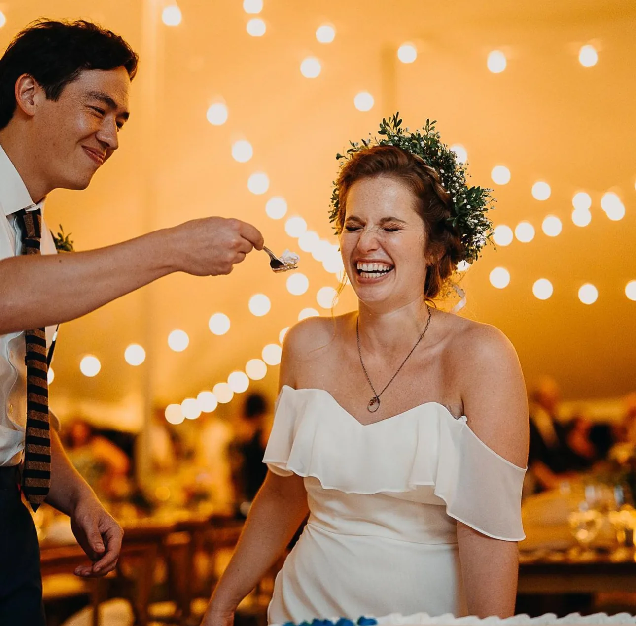 Bride laughing as her groom offers her a bite of cake under string lights