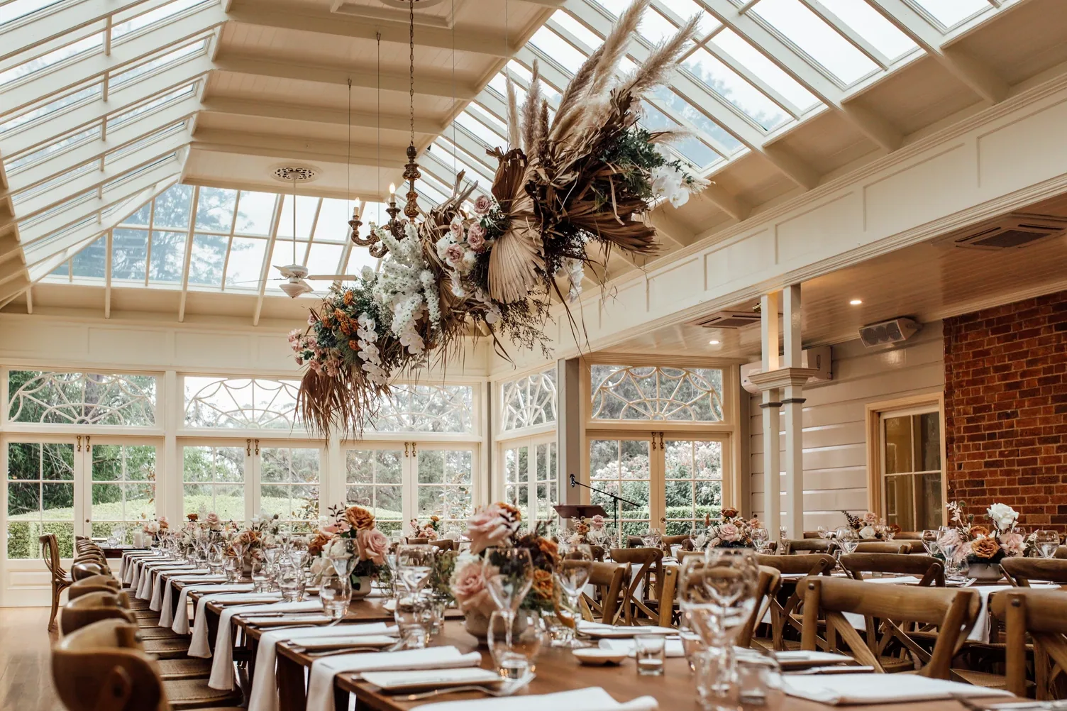 Long reception tables set beneath a glass roof with a hanging dried floral installation